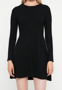 Femme portant une robe noire en maille côtelée à manches longues, taille cintrée et jupe évasée, debout devant un fond uni.