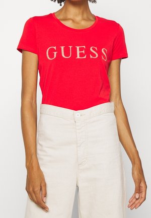 Femme portant un t-shirt rouge à manches courtes avec le mot « GUESS » en grandes lettres et un pantalon taille haute écru sur un fond uni.