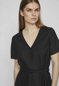 Juoda lino suknelė su trumpomis rankovėmis, V formos iškirpte, priekyje su sagų uždarymu ir surišamu juosmuo. Lygi tekstūra, minimalistiškas dizainas.