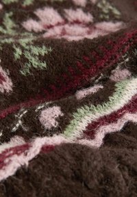 Close-up van zacht bruin gebreid stof met roze, groen en bordeauxrode bloemmotieven en gestructureerde stiksels.