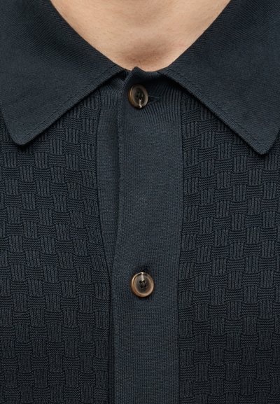 Κοντινή λήψη σκούρου ναυτικού πλεκτού πόλο με υφή, με γιακά τύπου spread και δύο καφέ κουμπιά κουμπωμένα στην κορυφή.