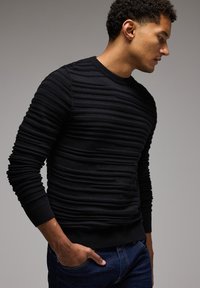 Schwarzer gestrickter Pullover mit strukturierten horizontalen Streifen, Rundhalsausschnitt und gerippten Bündchen. Kombiniert mit dunkelblauer Denim-Jeans, die eine figurbetonte Silhouette zeigt.