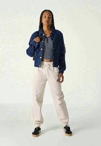 Una giovane donna sta in piedi indossando una giacca blu scuro, un top corto a righe bianche e nere, pantaloni felpati beige e sneakers nere con strisce bianche.