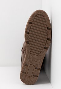 Brązowa podeszwa buta z teksturowanym i żłobionym bieżnikiem, rozmiar 37, umieszczona pionowo przy białych narożnych ścianach.