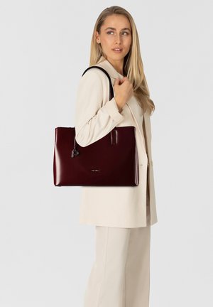 Bolso tote de cuero burdeos con acabado brillante, con asas largas y herrajes mínimos, sostenido por una persona con un conjunto crema.