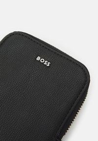 Portafoglio in pelle nera con superficie testurizzata, caratterizzato da un logo "BOSS" argentato e una cerniera dorata lungo il bordo.