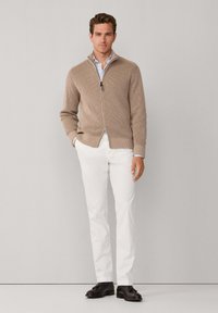 Beige gebreide trui met rits en ribbelstructuur, gecombineerd met witte broek en donkere schoenen. De trui heeft een hoge kraag en een gebreid ontwerp.