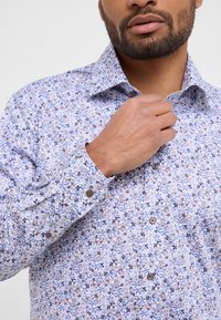 Lichtblauw overhemd met bloemenpatroon en paarse en beige accenten. Beschikt over een klassieke kraag, lange mouwen en knoopsluitingen.
