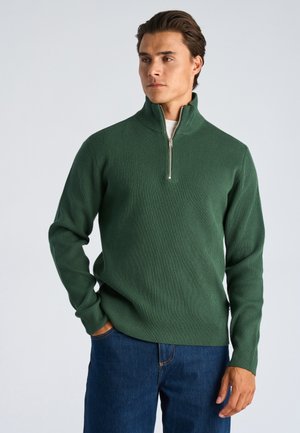 Homme portant un pull vert foncé côtelé avec zip quart, une chemise blanche en dessous et un jean bleu, debout avec une main dans la poche sur un fond uni.