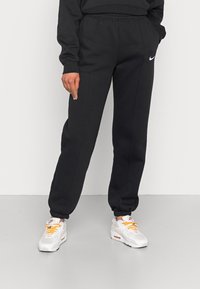 Black sweatpants with an elastic waistband and cuffs, featuring a white Nike logo on the front, worn with white sneakers and orange laces.
