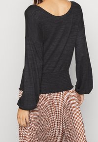 Black long-sleeve top with a relaxed fit, featuring a smocked waist and wide sleeves. Paired with a patterned, pleated skirt in brown and white.
