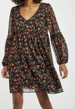 Kvinde iført en sort blomsterkjole med V-hals og gennemsigtige, lange puffærmer, prydet med små lyserøde, orange og gule blomster.