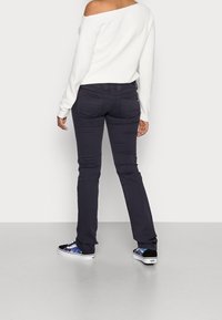Persoon in een off-shoulder wit trui, donkere strakke broek en zwart-witte sneakers met een kleurrijk patroon, staand tegen een effen muur.