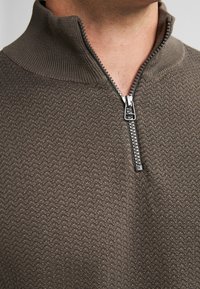Maglione marrone con zip, dalla superficie texturizzata e colletto alto. Presenta una zip metallica con un taglio a strappo, che mostra un motivo a zigzag.