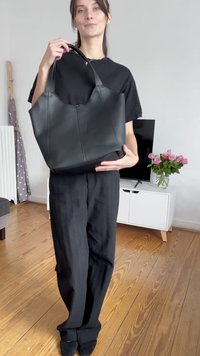 Femme en tenue noire tenant un grand sac tote en cuir noir dans un salon avec des sols en bois et un meuble de télévision.