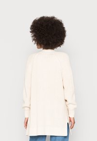 Vaaleanbeige neulecardigan, jossa on tekstuurinen kuvio, raglan-hihat ja sivuhalkiot liikkumisen helpottamiseksi. Ribbaatut hihansuut ja helman yksityiskohtia.