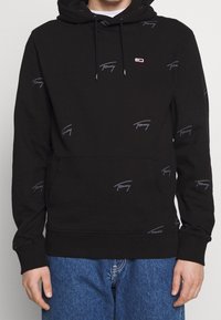 Sudadera negra de algodón con una textura suave, que presenta un patrón de logo repetido en gris y un bolsillo delantero. Incluye cordones ajustables.