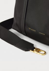 Mustanharmaa kangaspussi, jossa on kultainen metallilukko, sileä pinta, säädettävä hihna ja kohokuvioitu brändilogo.