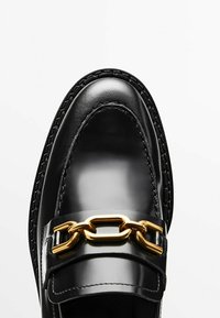 Mocassin en cuir noir avec une finition brillante, orné d'une chaîne dorée et de coutures tressées le long de la semelle. Texture lisse sur toute la surface.
