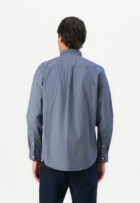 Hombre de pie con la espalda hacia la cámara, vistiendo una camisa azul de manga larga con un diseño y pantalones oscuros, contra un fondo blanco liso.
