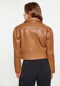 Giacca cropped in finta pelle marrone con maniche a sbuffo, design su misura e superficie liscia. Completata con colletto classico e cuciture posteriori.