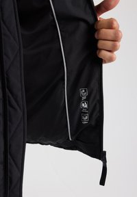 Black padded jacket interior features windproof and water-repellent labels, contrasting white piping, and a zippered pocket at the hem.