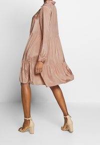 Donna che indossa un vestito lungo, a maniche lunghe, di un rosa polveroso fino al ginocchio e sandali con tacco alto beige con cinturino alla caviglia, mentre cammina.