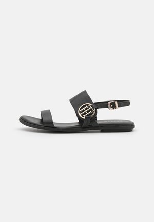 Sort flad sandal med to stropper, en guldfarvet rund logodetalje og en justerbar ankelspænde på en hvid baggrund.
