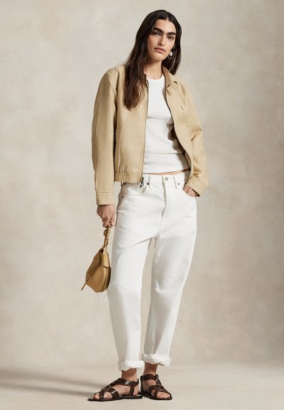 Beige bomberjakke, hvit ribbestrikket tanktopp, hvite avslappede jeans og brune sandaler, holder en liten tan-farget håndveske med vridd stropp.