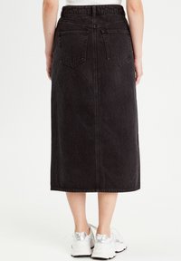 Jupe midi en denim noir avec une coupe droite, dotée de poches arrière et d'une texture lisse. Portée avec des chaussures de sport blanches et argentées.