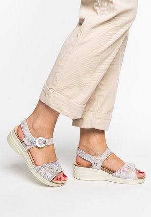 Feet wearing floral-patterned wedge sandals with ankle straps and beige rolled-up pants against a white background.