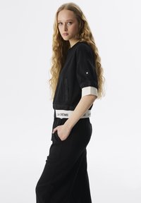 Schwarzes Crop-Top mit Rundhalsausschnitt, kurzen Ärmeln und einem weißen Band an der Taille mit dem Text "LA CRÈME". Kombiniert mit schwarzen Hosen.