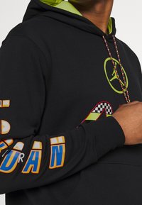 Svart hoodie med en grön logotyp, livfull flerfärgad text och rutiga mönsterdetaljer. Dragsko med texturerade, flerfärgade fibrer.