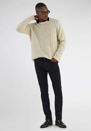 Mann trägt cremefarbenen Zopfpullover, schwarze Jeans, schwarze Stiefel und Sonnenbrille, posiert mit einer Hand am Nacken und der anderen in der Tasche.