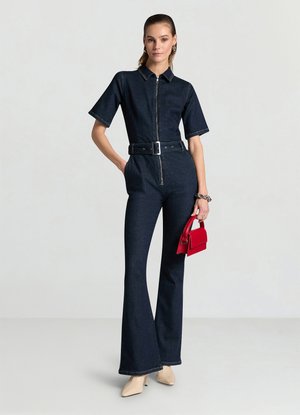 Tuta in denim scuro con maniche corte, zip frontale, vita cintata, gambe svasate e colletto standard. Abbinata a una piccola borsa rossa.