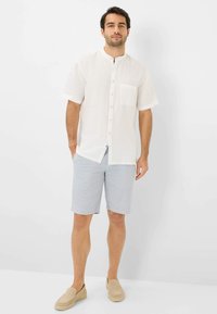 Camicia bianca di lino a maniche corte con taschino sul petto, abbinata a pantaloni corti a strisce blu chiaro. Scarpe slip-on color sabbia. Sfondo neutro.