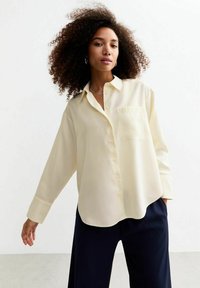 Crème-kleurige button-up shirt van een gladde stof, met een borstzak en lange mouwen, gecombineerd met donkere broeken.