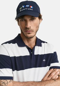 Marinblå keps med "GANT USA" broderat, med färgglada flaggor. Modellen bär en marinblå och vitrandig polo med krage.