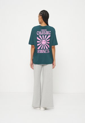 Άτομο με πλεξούδες που στέκεται με την πλάτη, φοράει μια μπλούζα τυρκουάζ με ροζ γραφικό ήλιου και το κείμενο «Forever Chasing Sunsets» και ανοιχτό γκρι φαρδιά παντελόνια.