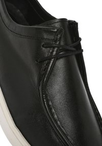 Chaussure en cuir noir avec une texture lisse, bout arrondi et semelle en caoutchouc blanche contrastante. Les lacets noirs et le détail de couture sont visibles.