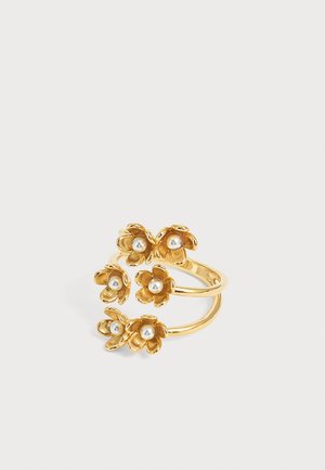 Bague ton or avec trois anneaux ouverts, chacun orné de petites formes florales tenant des perles blanches au centre, sur un fond neutre.