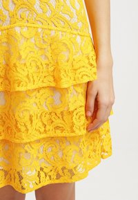 Main reposant à côté d'une jupe en dentelle jaune à motifs floraux et volants superposés sur un fond clair.