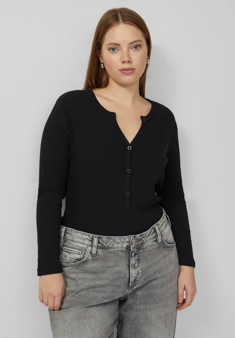 Zwart geribd langärmelig topje met een V-hals, voorzien van een knoopsluiting. Gecombineerd met lichtgrijze, losse spijkerbroek.