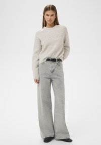 Žena v světle šedém chlupatém svetru zastrčeném do kalhot s vysokým pasem v acid-wash střihu a širokých nohavicích, s černým páskem a černými špičatými botami.
