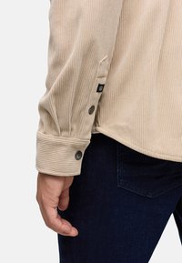 Camisa de pana beige con textura acanalada, puños con botones de metal y una pequeña etiqueta negra en la manga, combinada con jeans azul oscuro.