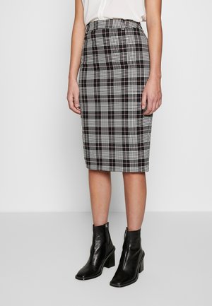 Vrouw die een knielange zwart-wit geruite kokerrok draagt met zwarte enkellaarzen en een witte blouse tegen een effen achtergrond.