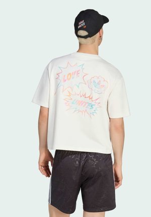 Personne portant un T-shirt blanc avec un texte et un graphique pastel « LOVE UNITES », un short noir avec des motifs subtils et une casquette noire assortie.