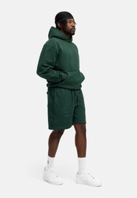 Grüne Kapuzenjacke mit einer Vordertasche und passenden Shorts, beide aus strukturierter Stoff gefertigt. Kombiniert mit weißen Sneakern und Socken in Crew-Länge.