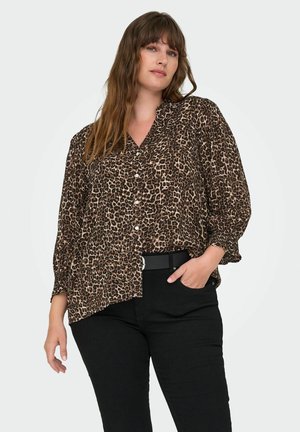 Femme aux longs cheveux bruns portant une blouse à motifs léopard et un pantalon noir avec une ceinture noire, posant avec une main dans la poche.