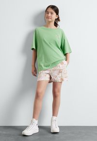 Žalia medvilninė didelio dydžio trumpa rankovė T-shirt, derinama su raštuotais rausvais ir smėlio spalvos šortais. Modelis vilki baltas aukštakulnes sportines batelius.